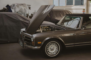 Classic Mercedes-Benz W113 Pagoda with its bonnet open in a restoration workshop, alongside other covered classic cars
