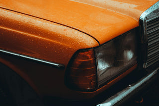 Close-up of a classic Mercedes-Benz front corner, headlight and indicator lens, with rain droplets on the orange bodywork